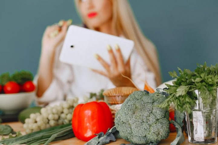 Blonde woman using tablet with fresh vegetables like broccoli and tomatoes in a kitchen setting.